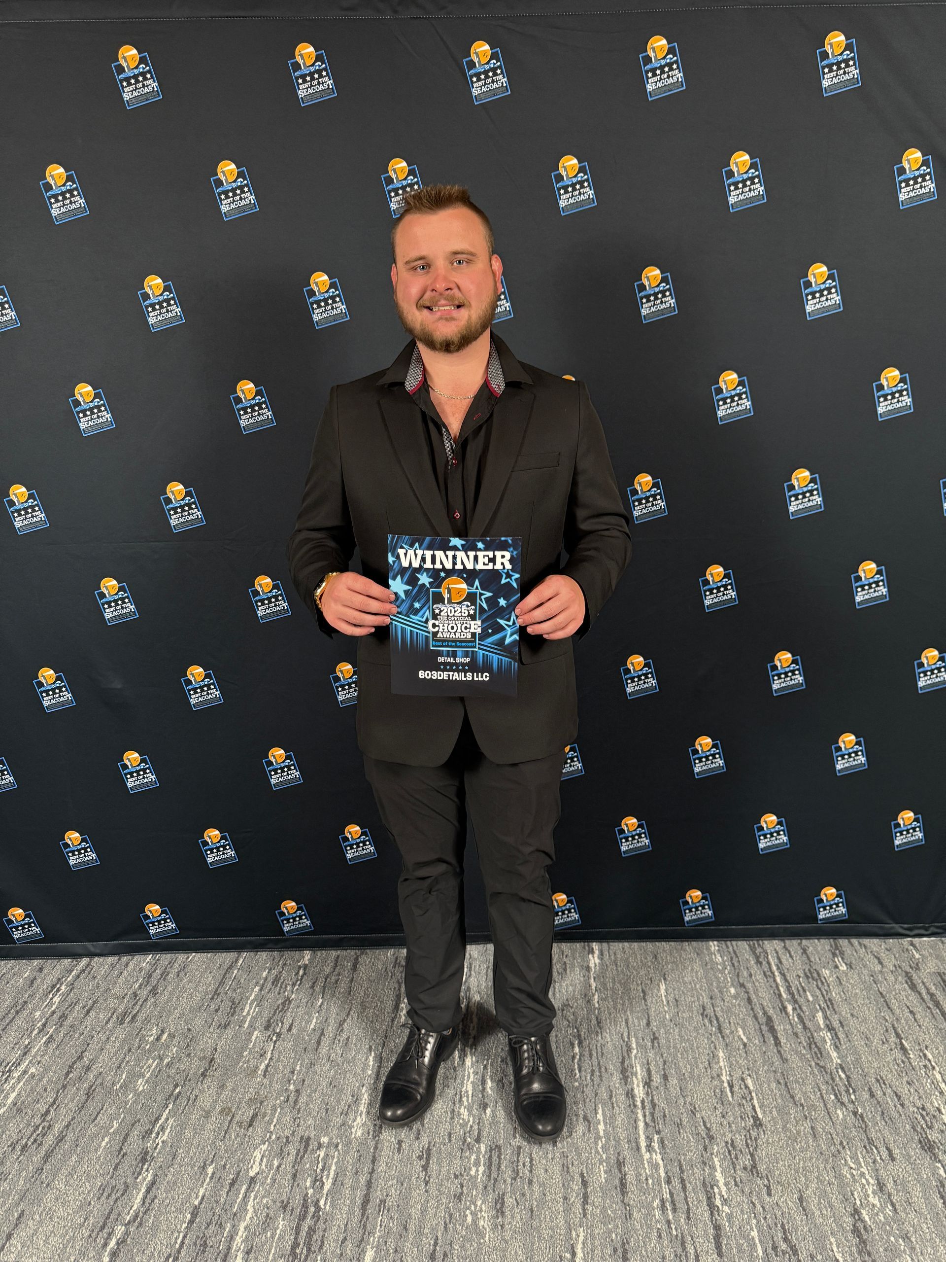 Man standing before a branded backdrop, holding a book or magazine and smiling at the camera.