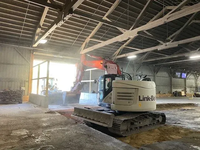 Excavator inside a warehouse, with a bucket scooping material near an open doorway.