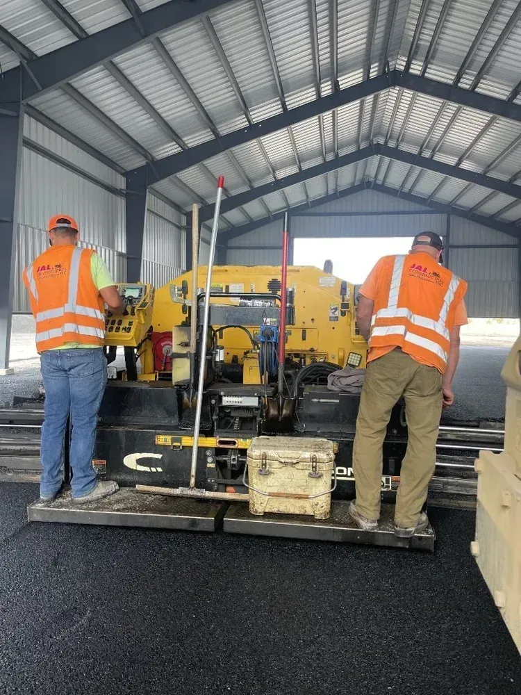 Workers in orange vests operating a yellow paving machine inside a metal-roofed shed