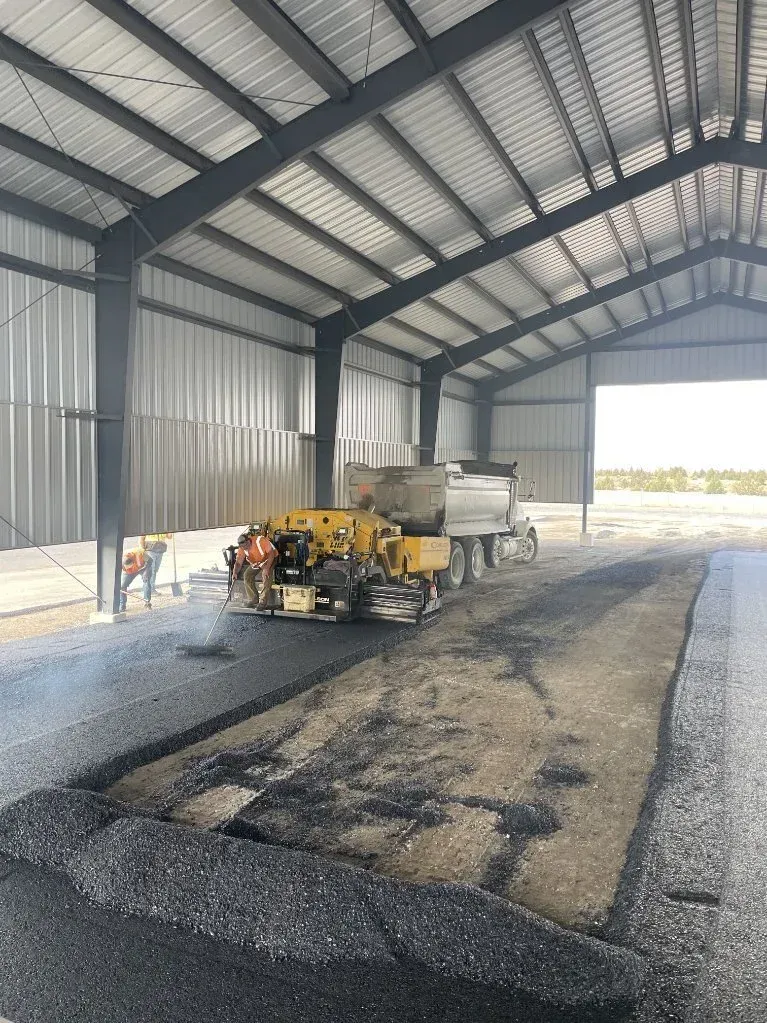 Industrial shed with a yellow machine on a gravel floor and daylight coming through the open end.
