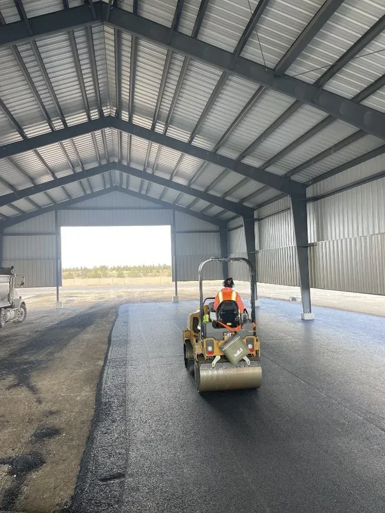 Paving machine laying asphalt inside a metal-roofed shed, with a worker seated on it.