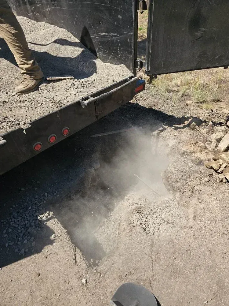 Dust billows from a trailer unloading dirt onto a gravel surface.
