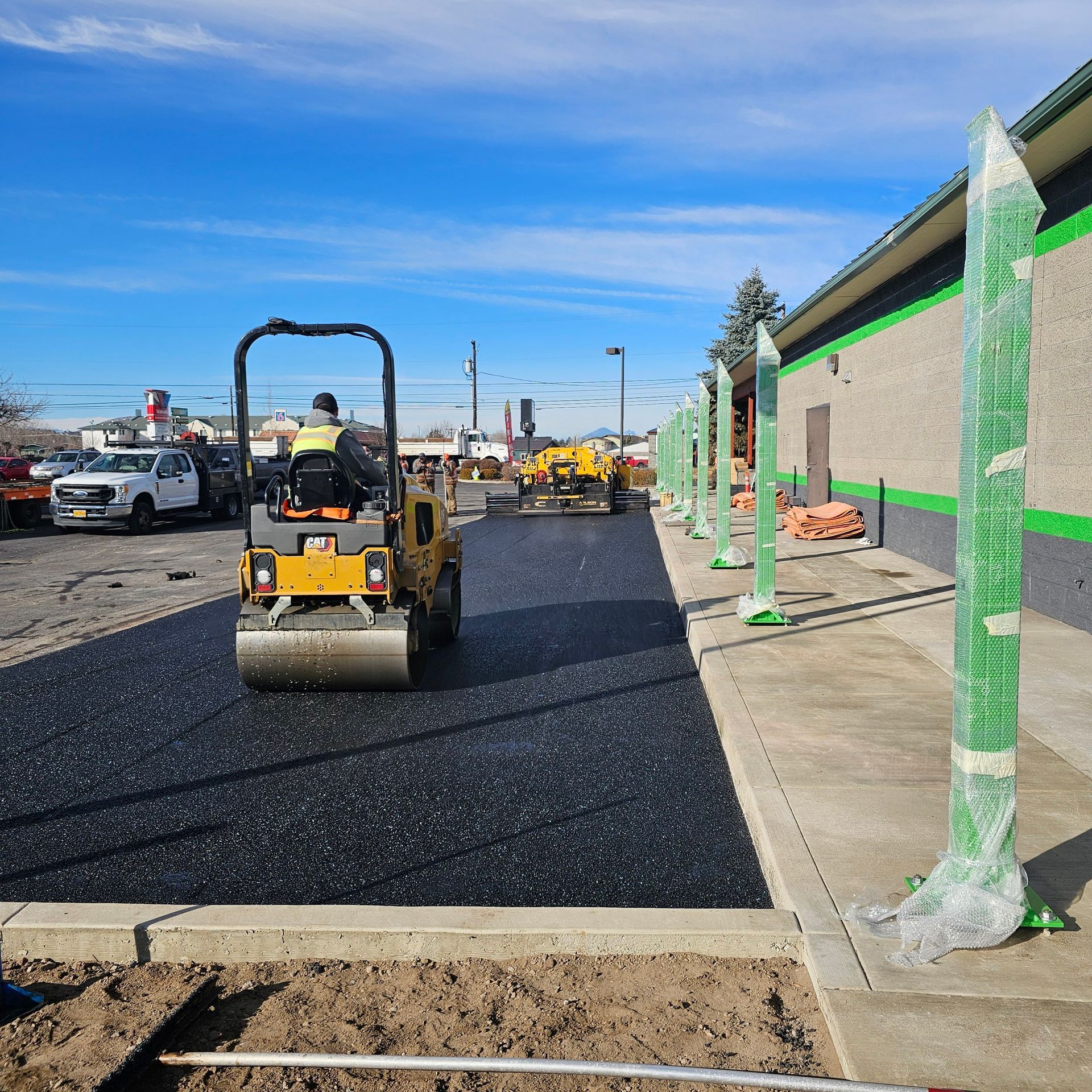 Road roller paving a parking lot beside a building with green-wrapped posts on a sunny day