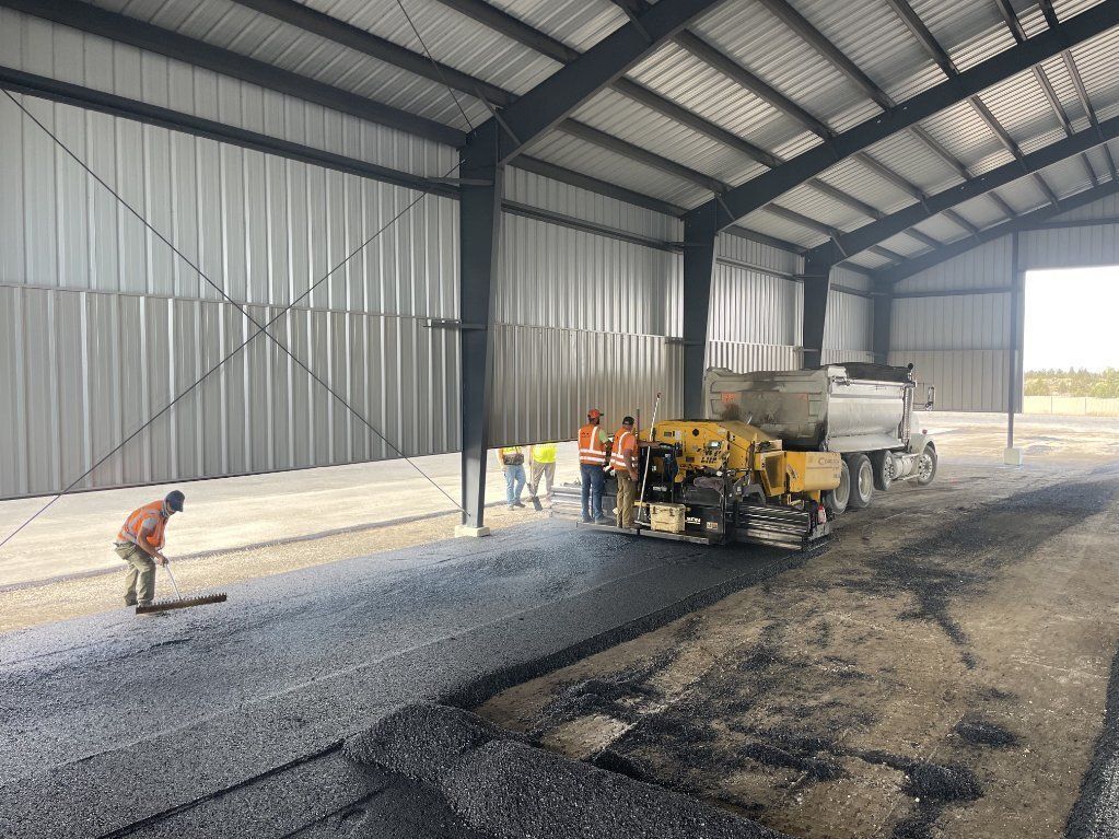 Worker operating paving machine inside metal warehouse, laying fresh asphalt beside a red-clad worker.
