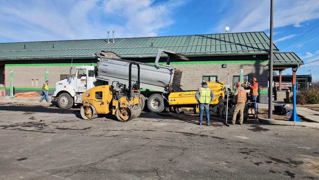 Workers beside a yellow loader and dump truck at a construction site near a green-roofed building