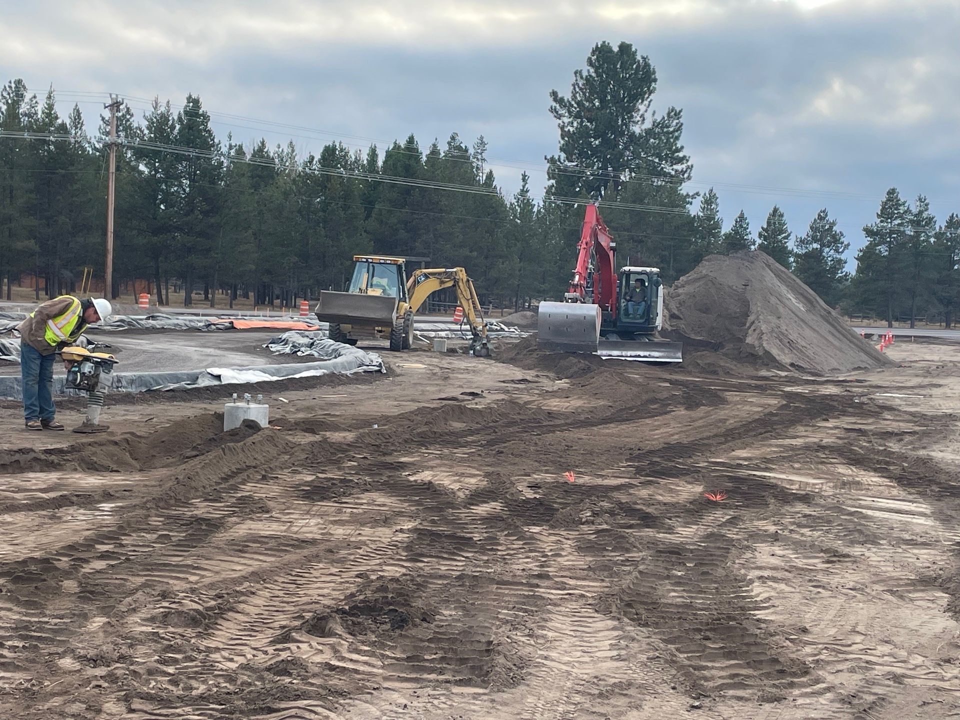 Construction site with excavator, red crane, dirt piles, and a worker in a wooded area under cloudy skies