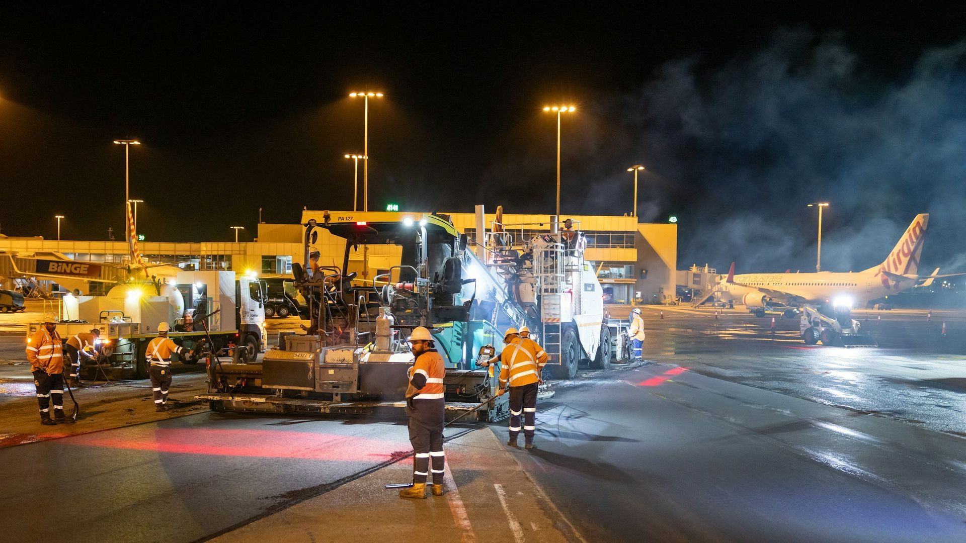 Ground crew on a lit airport tarmac near aircraft, with smoke and reflective wet pavement at night