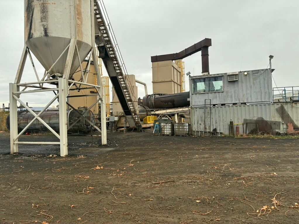 Industrial yard with silos, conveyor belts, and a corrugated metal building on a gravel lot under cloudy skies