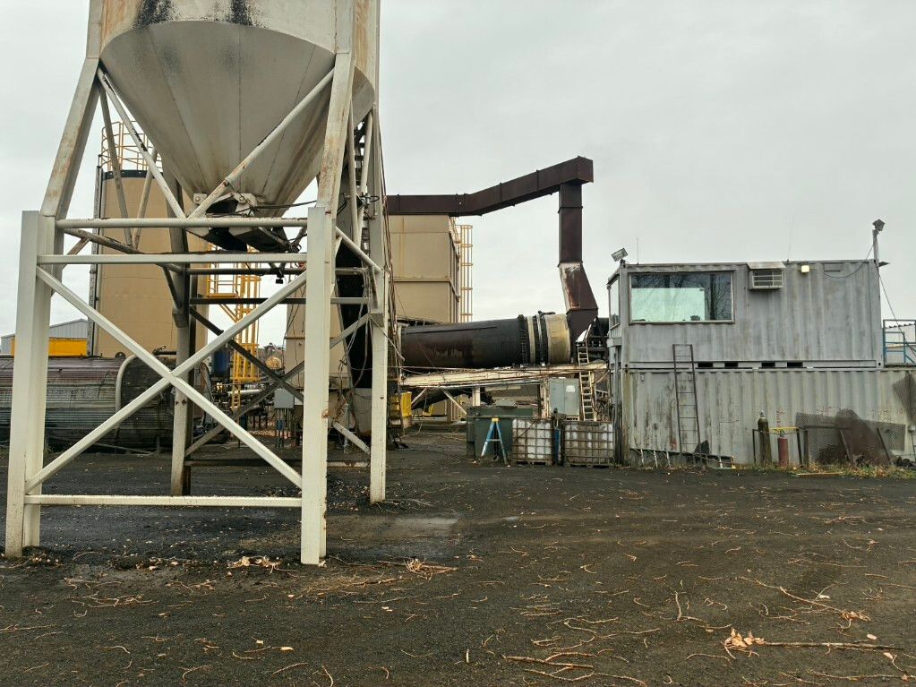 Industrial site with large metal hopper, conveyor, and weathered buildings under a gray sky