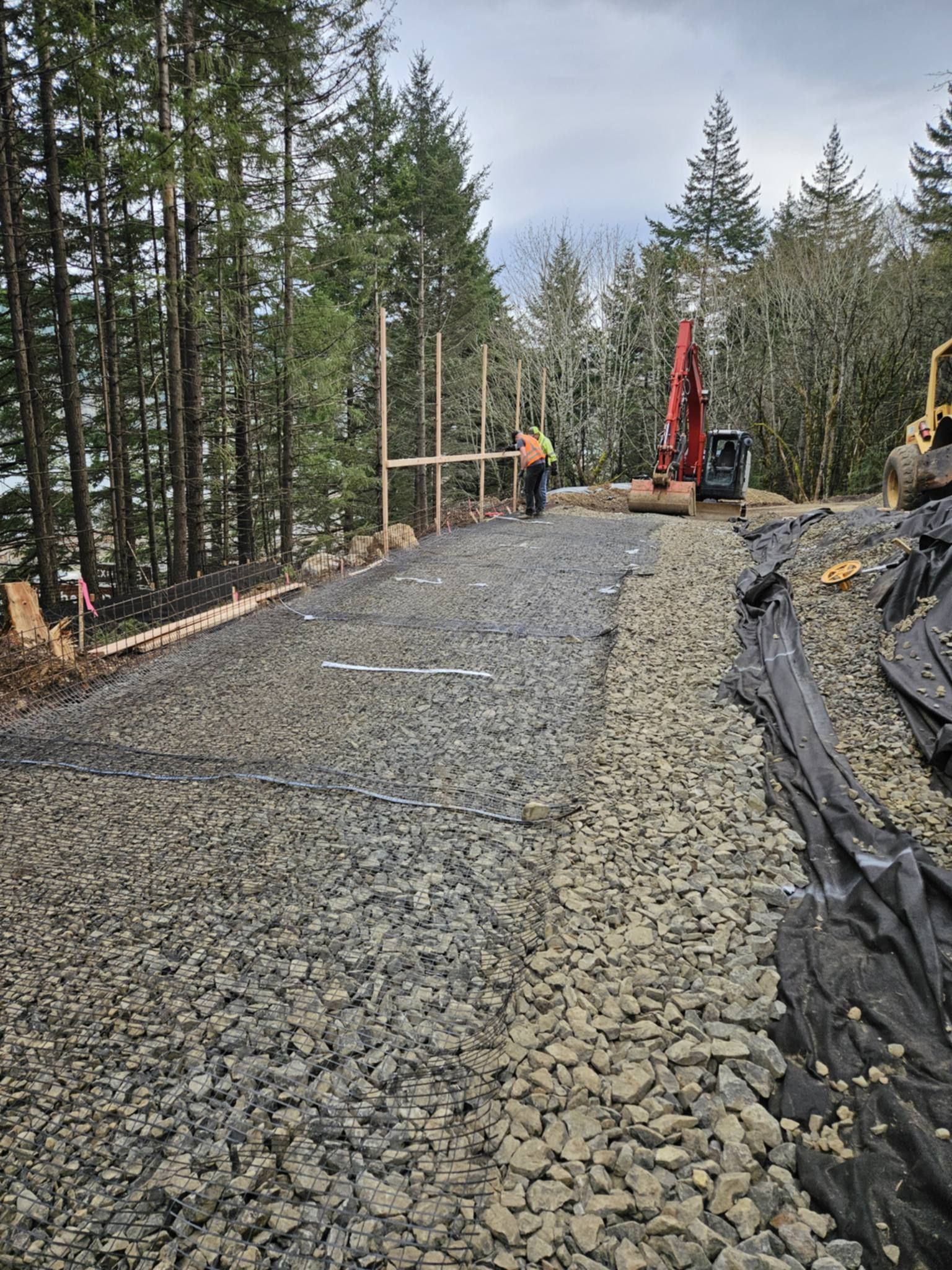 Gravel road under construction beside tall pine trees, with excavator and worker in the distance