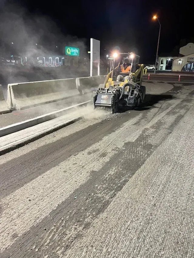 Roadwork at night with a skid-steer clearing debris beside a concrete barrier on a dusty road.