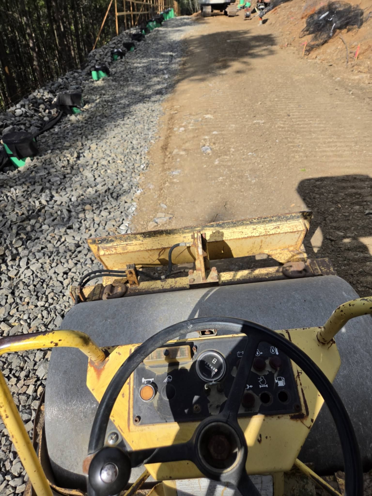 View from a road roller over a gravel road under construction, with a dirt shoulder and rocks along the left edge