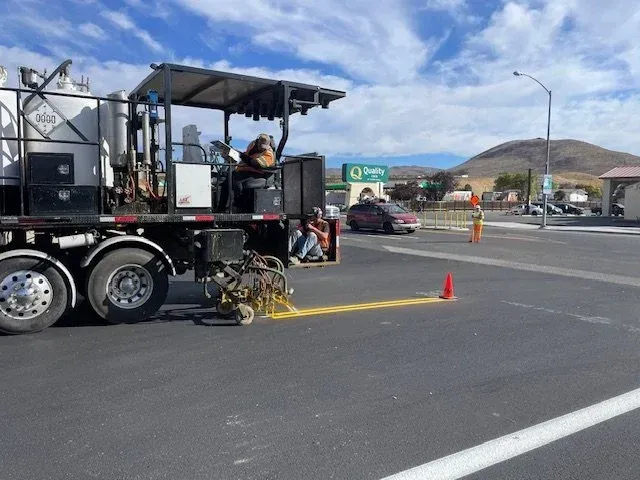 Road construction vehicle spraying yellow paint on asphalt, with cones and a worker nearby at an intersection.