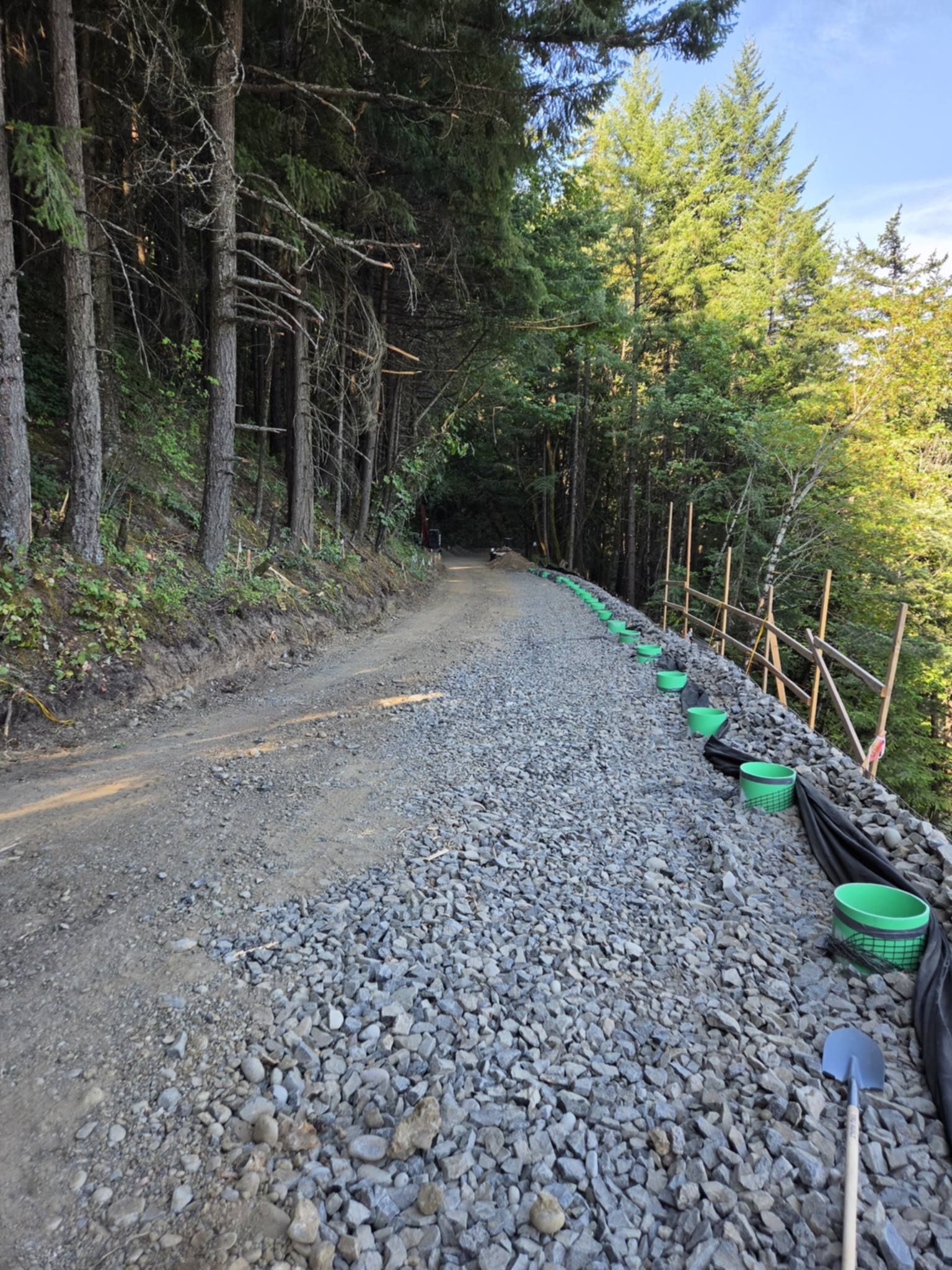 Gravel road under construction beside a forest, with green stakes and black edging along the right side.