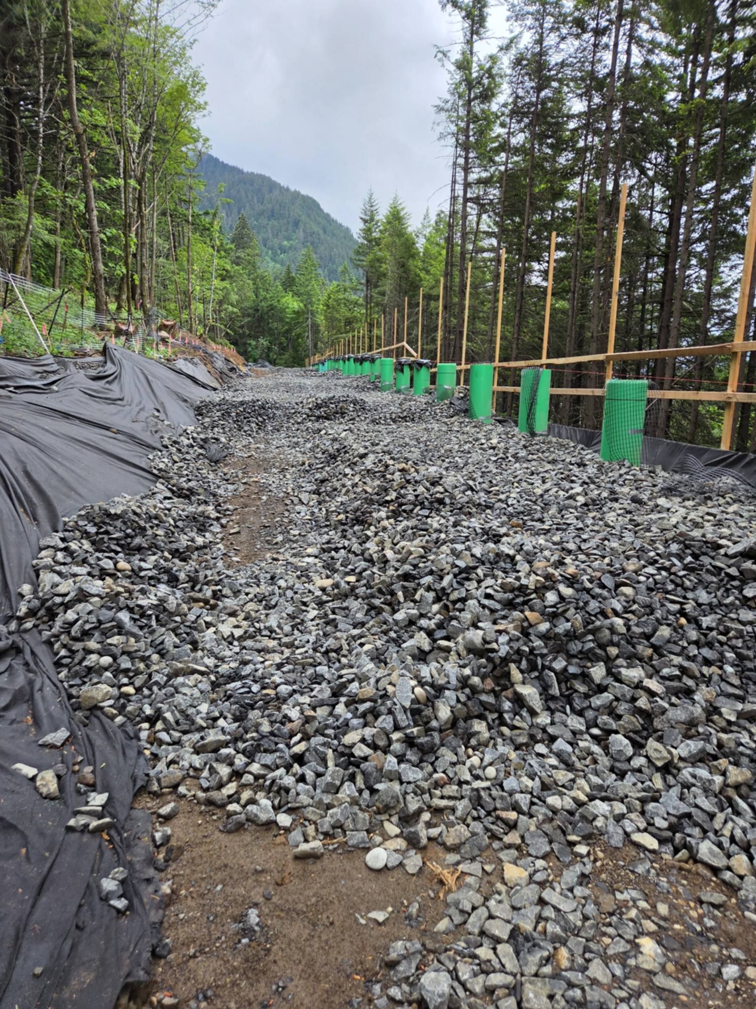 Rock embankment work on a forest road with tarps, green-wrapped posts, and mountains in the background