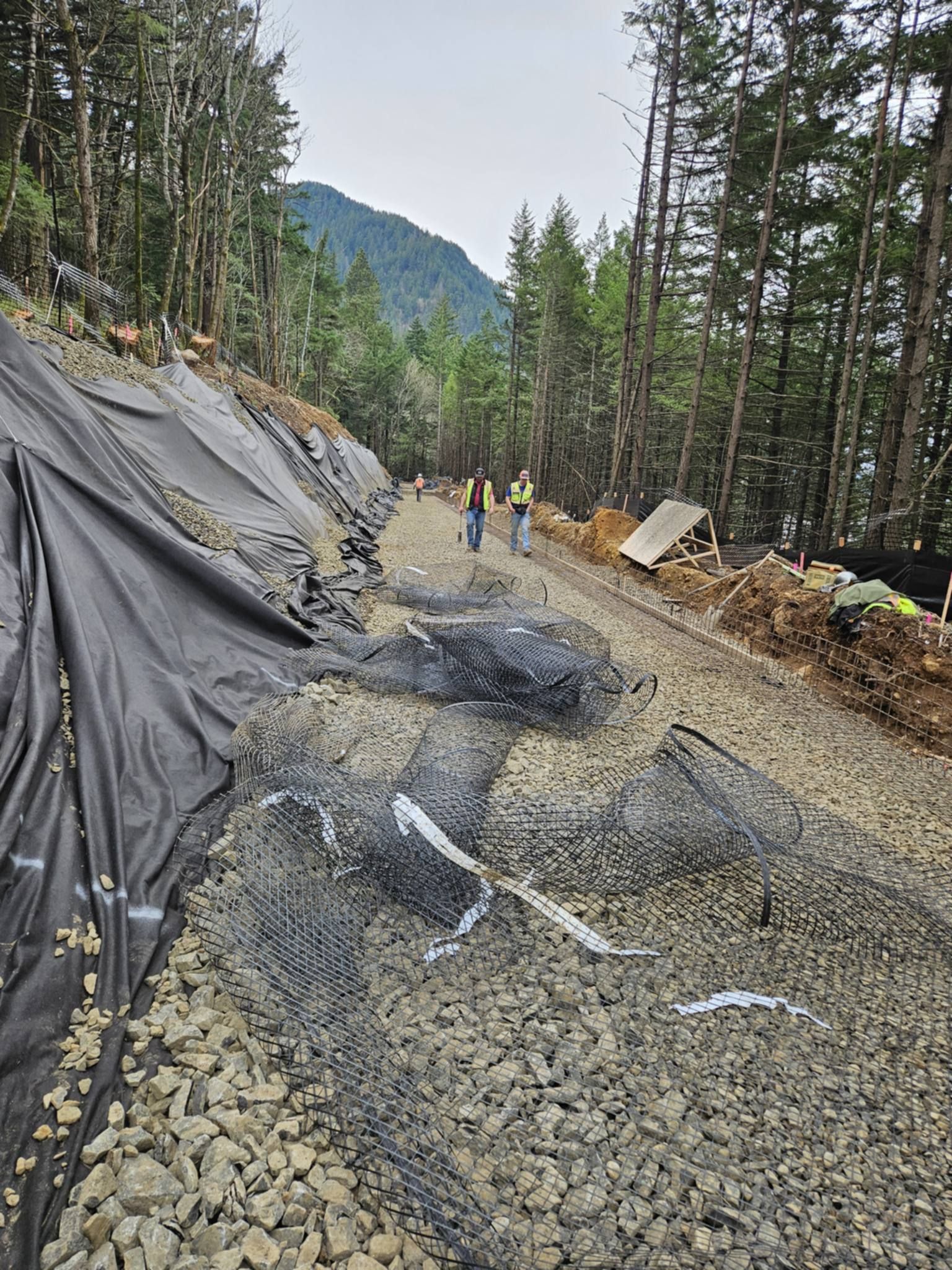 Collapsed mountain road with landslide debris, tarps, and workers on a gravel forest hillside