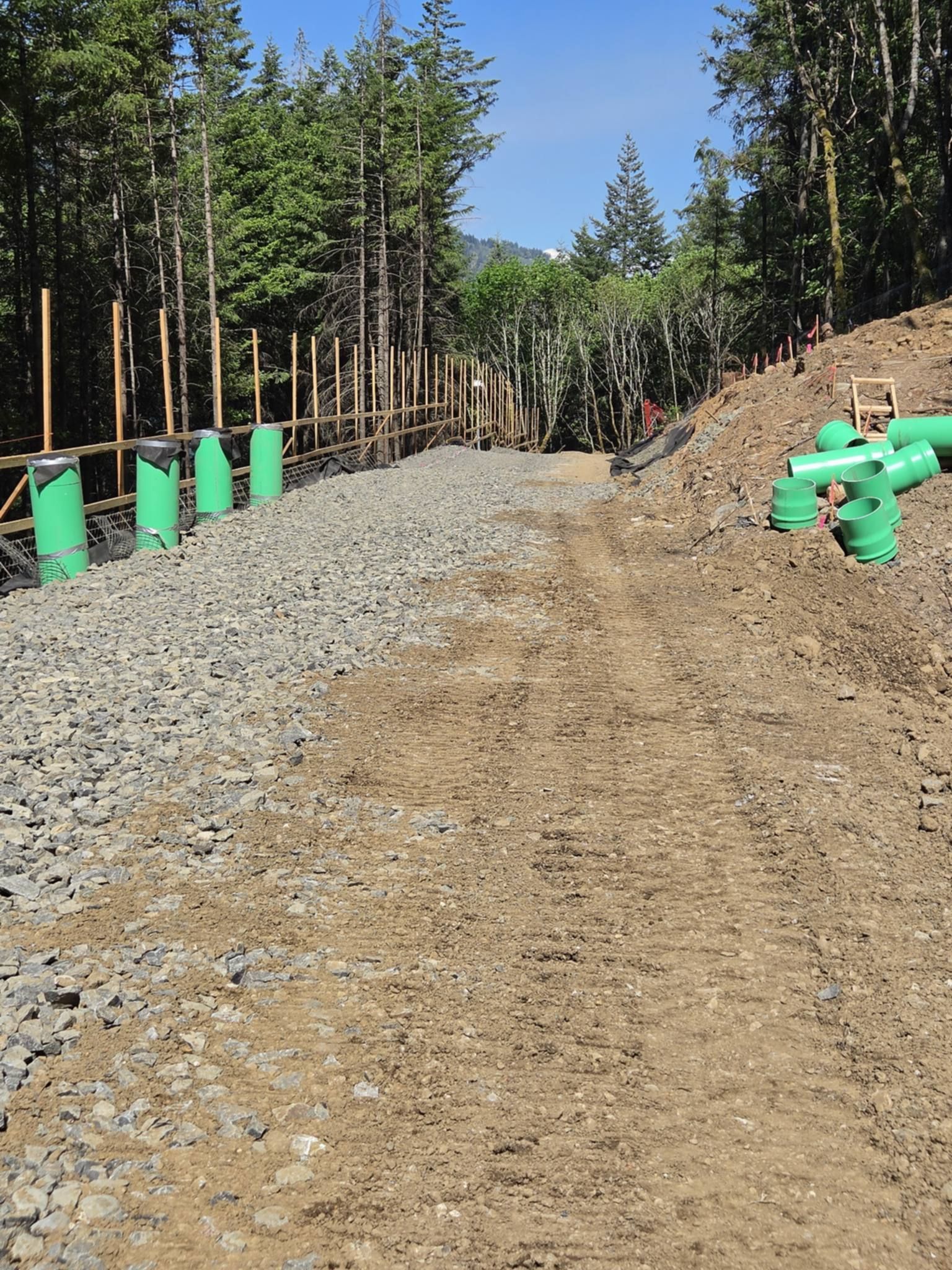 Gravel road under construction beside a dirt slope, with green utility pipes and trees lining both sides