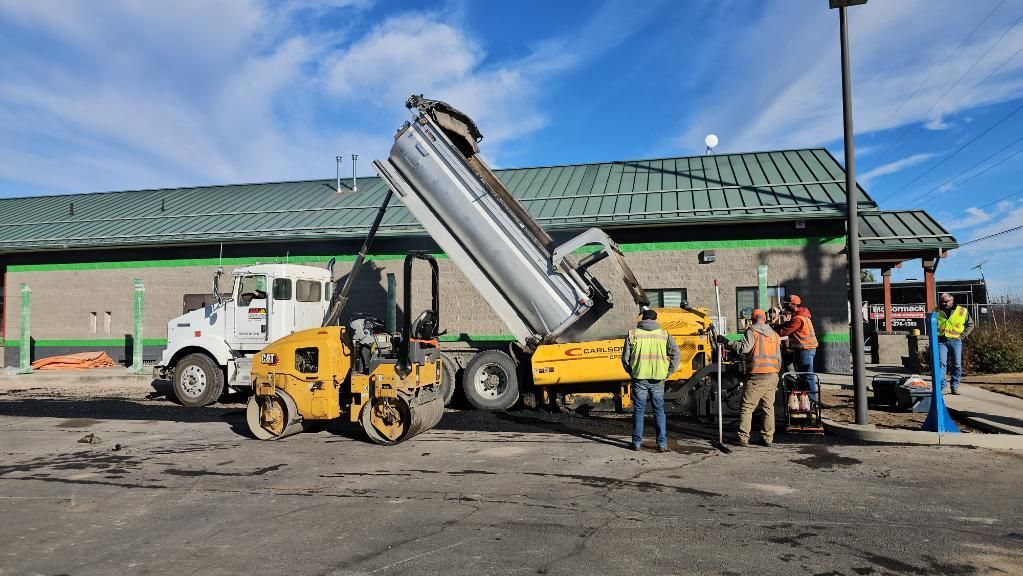 Workers and construction vehicles loading asphalt at a roadside work site under a green-roofed building.
