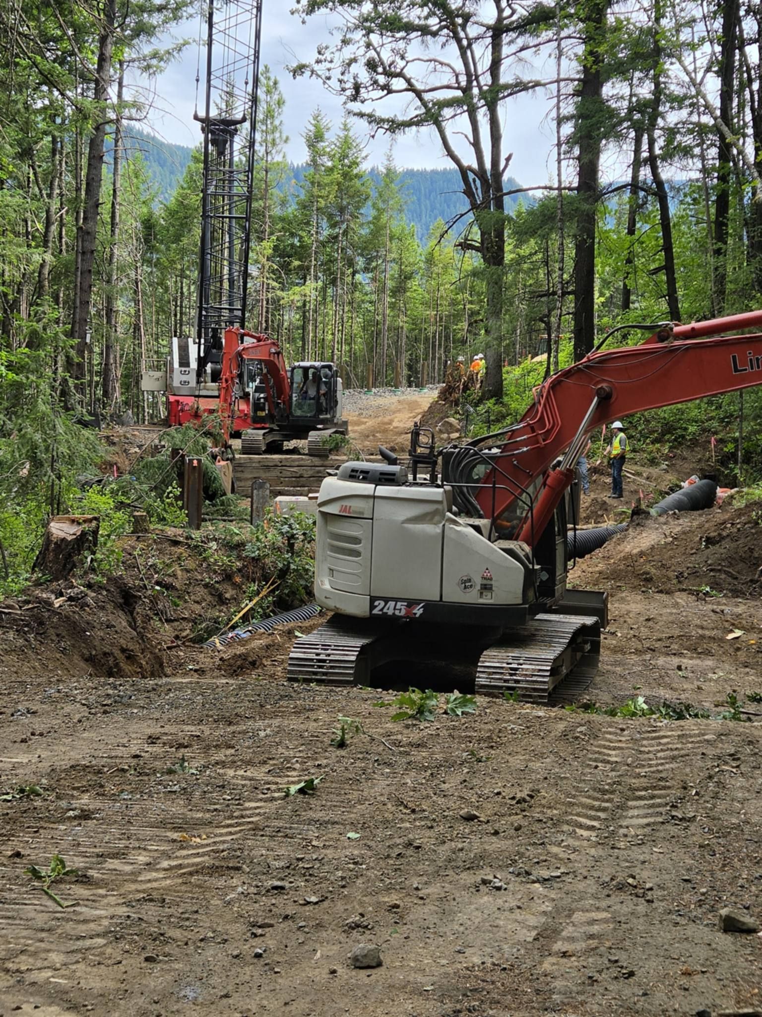 Two excavators on a muddy forest trail with one red arm in the foreground and trees in the background