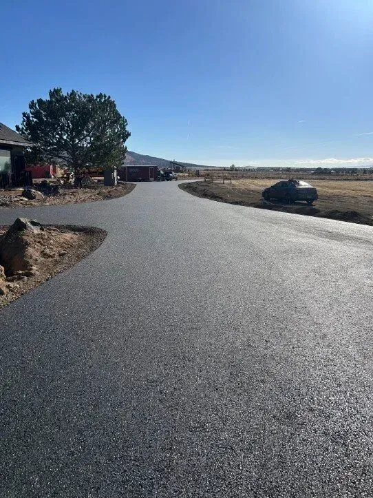 Newly paved rural road curving past a tree, houses, and open fields under a clear blue sky