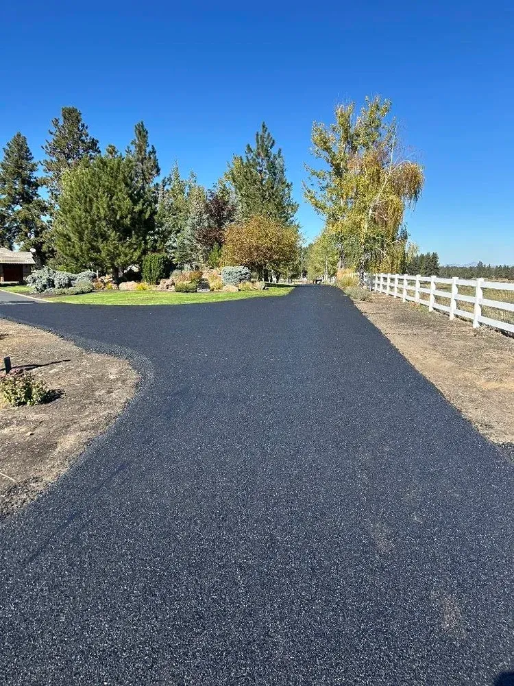 Fresh black asphalt driveway curving through a rural yard with trees and a white fence under blue sky