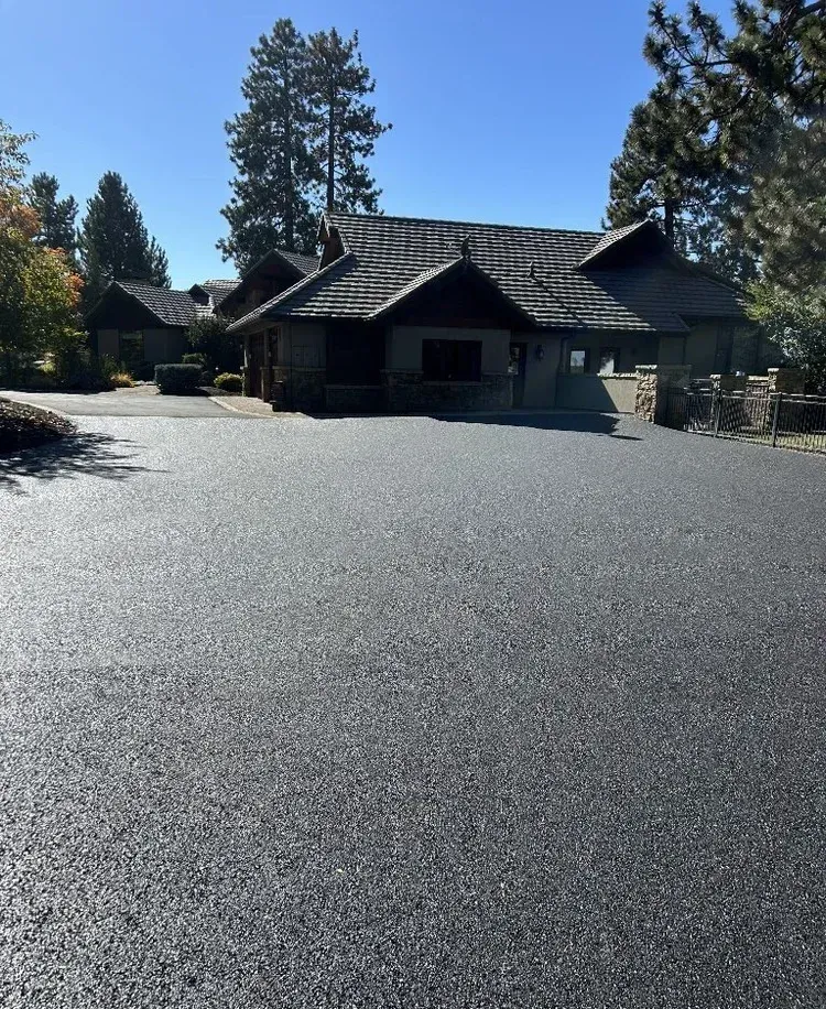 Large gravel driveway leading to a dark-roofed house with tall pine trees under a clear blue sky.