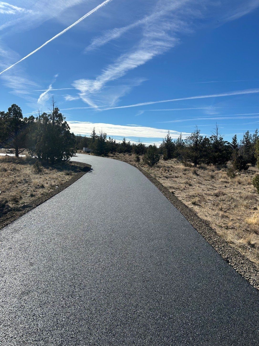 Curving paved path through dry grass and trees under a bright blue sky with contrails
