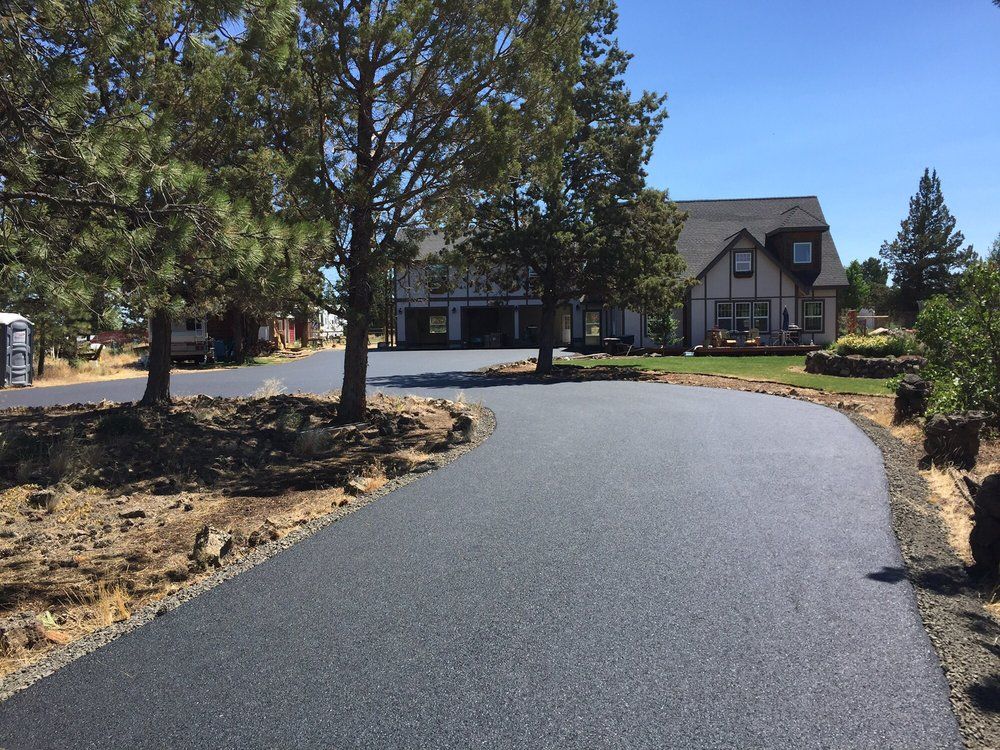Curved black asphalt driveway leading to a house with trees and landscaping on a sunny day