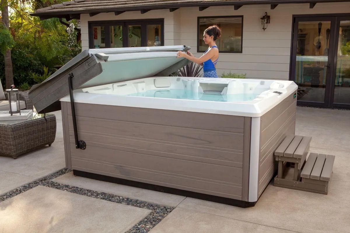 A woman is opening the lid of a hot tub on a patio.