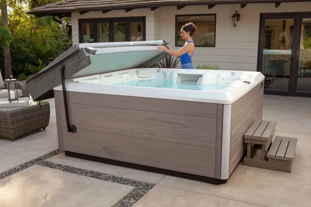 A woman is opening the lid of a hot tub on a patio.