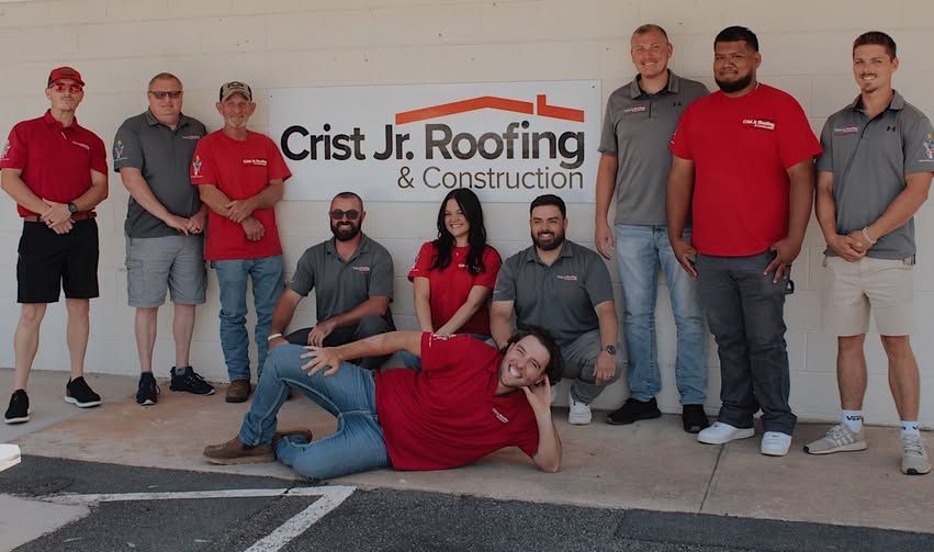 Group of employees posing in front of a Crist Jr Roofing & Construction sign, one man lying in front.