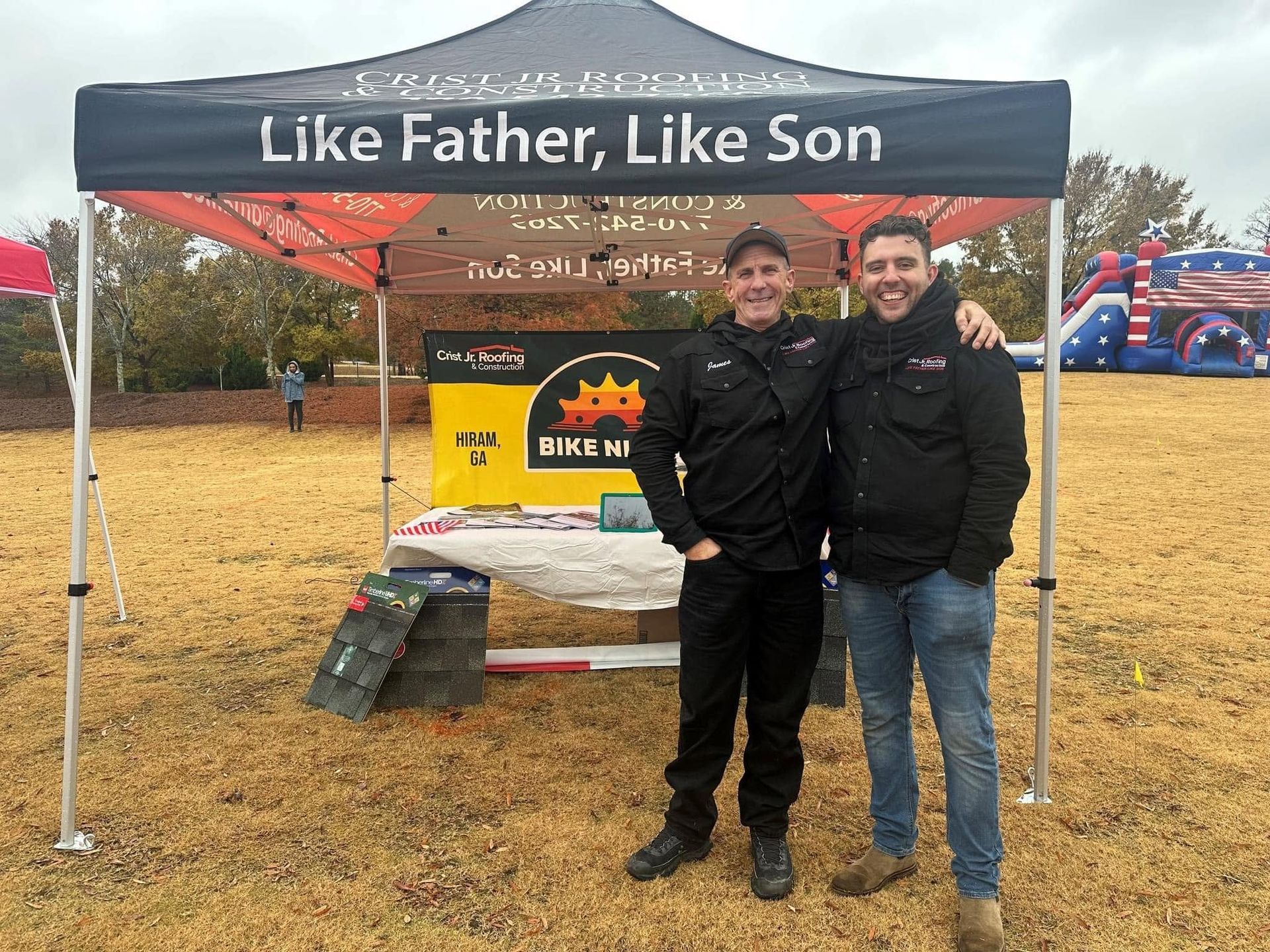 Two people standing under a tent with “Like Father, Like Son” banner at an outdoor event.