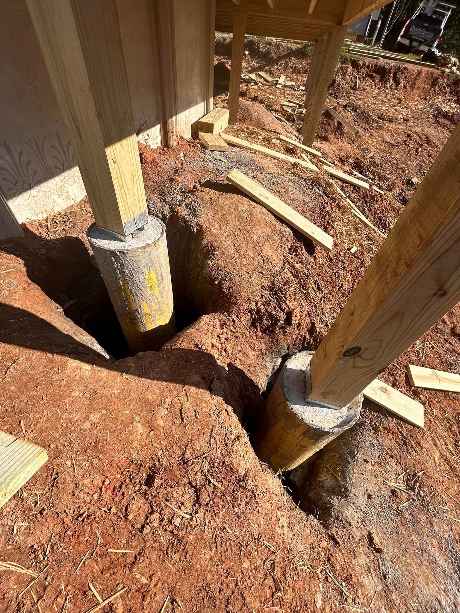 Wooden deck posts on concrete footings in a dirt excavation, with a hole and loose soil around the base