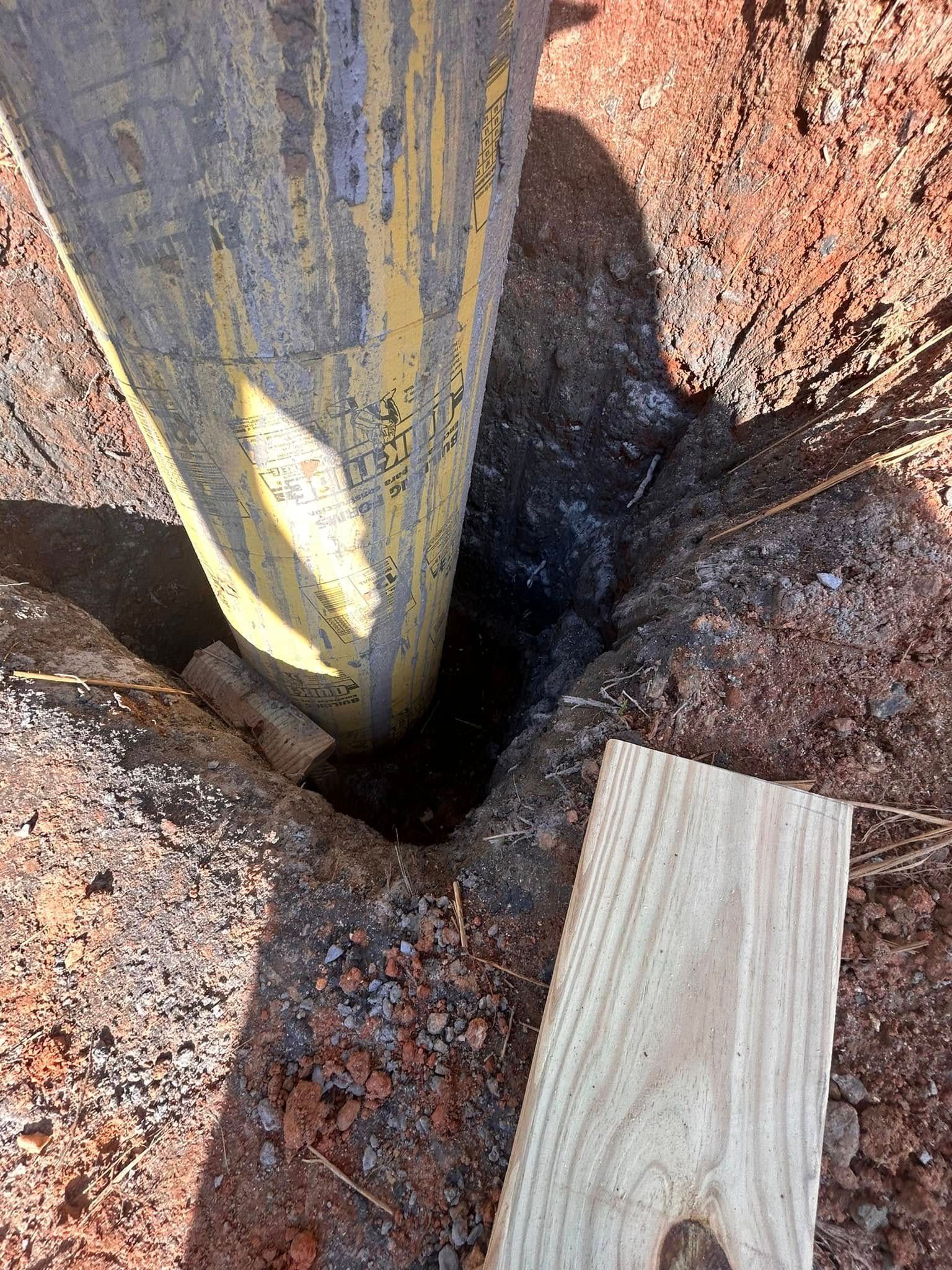 Wooden posts set in a small hole in reddish dirt, with a loose plank nearby.