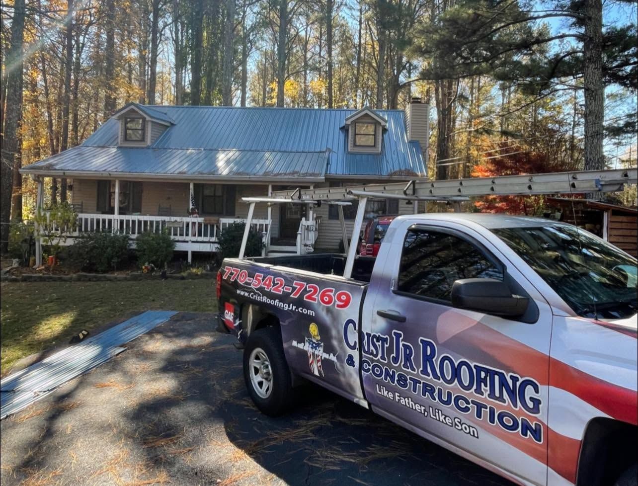 Roofing company truck parked in front of a house with a metal roof and trees in the yard