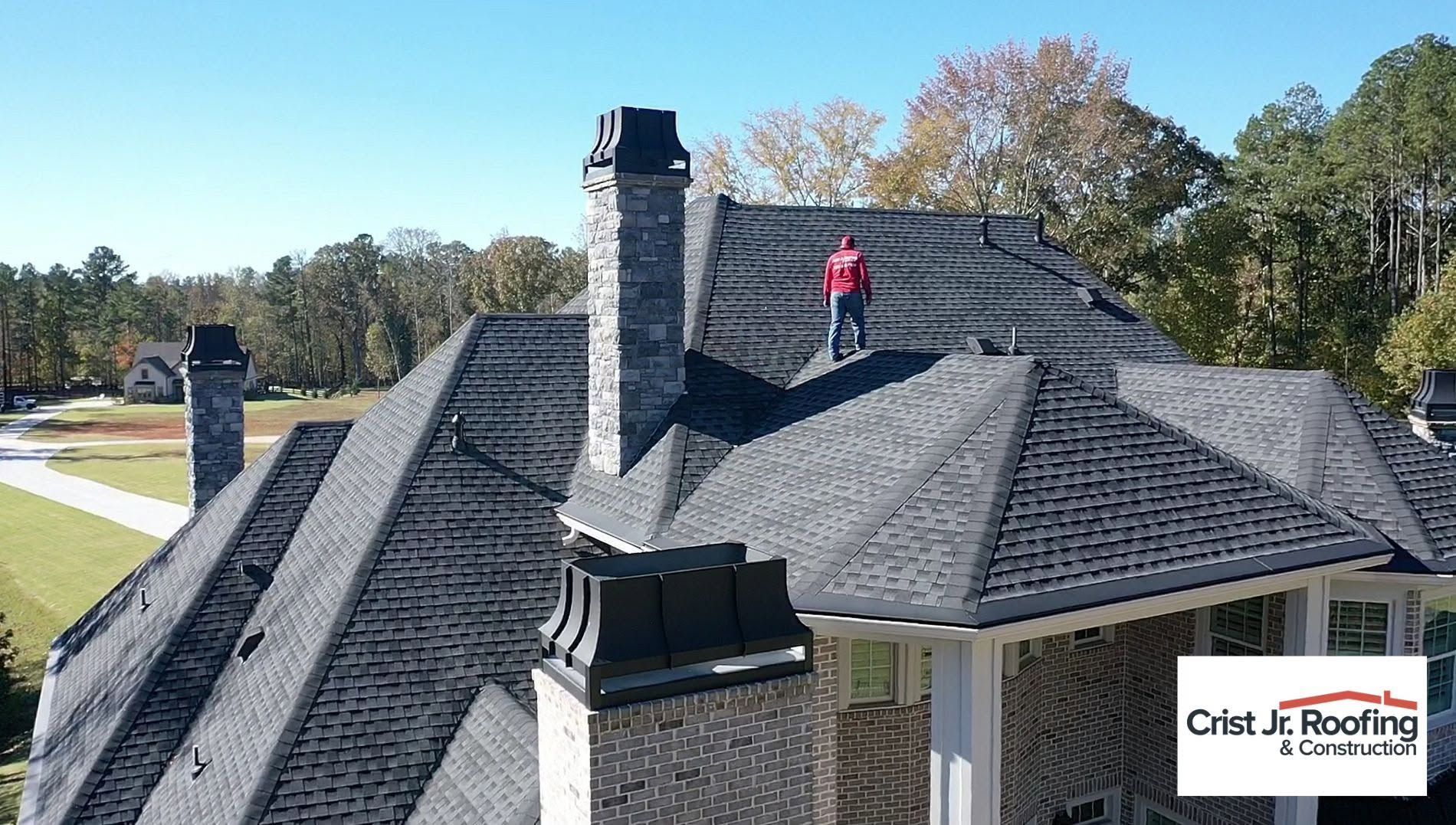 Roof inspection on a dark gray shingle roof, with a person in a red jacket near the chimney.