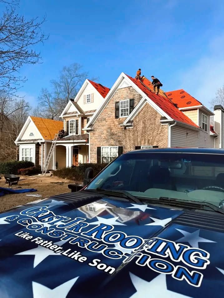 Roofers working on a brick house with red shingles, viewed past a black truck with a roofing company logo.