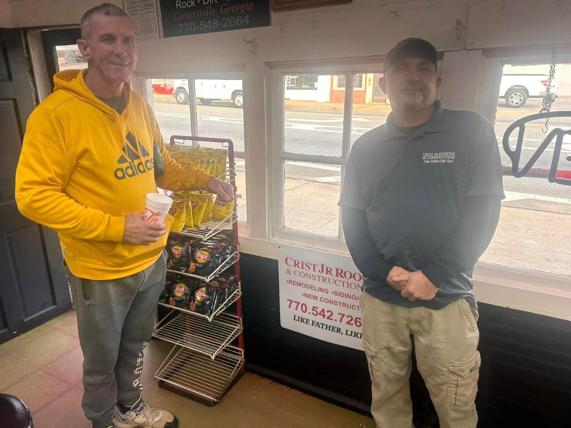Two men stand by a storefront window, one in a yellow hoodie holding a cup, the other in a dark sweatshirt.