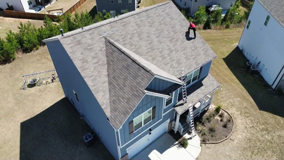 Aerial view of a gray house with a shingled roof and small front porch in a suburban yard.