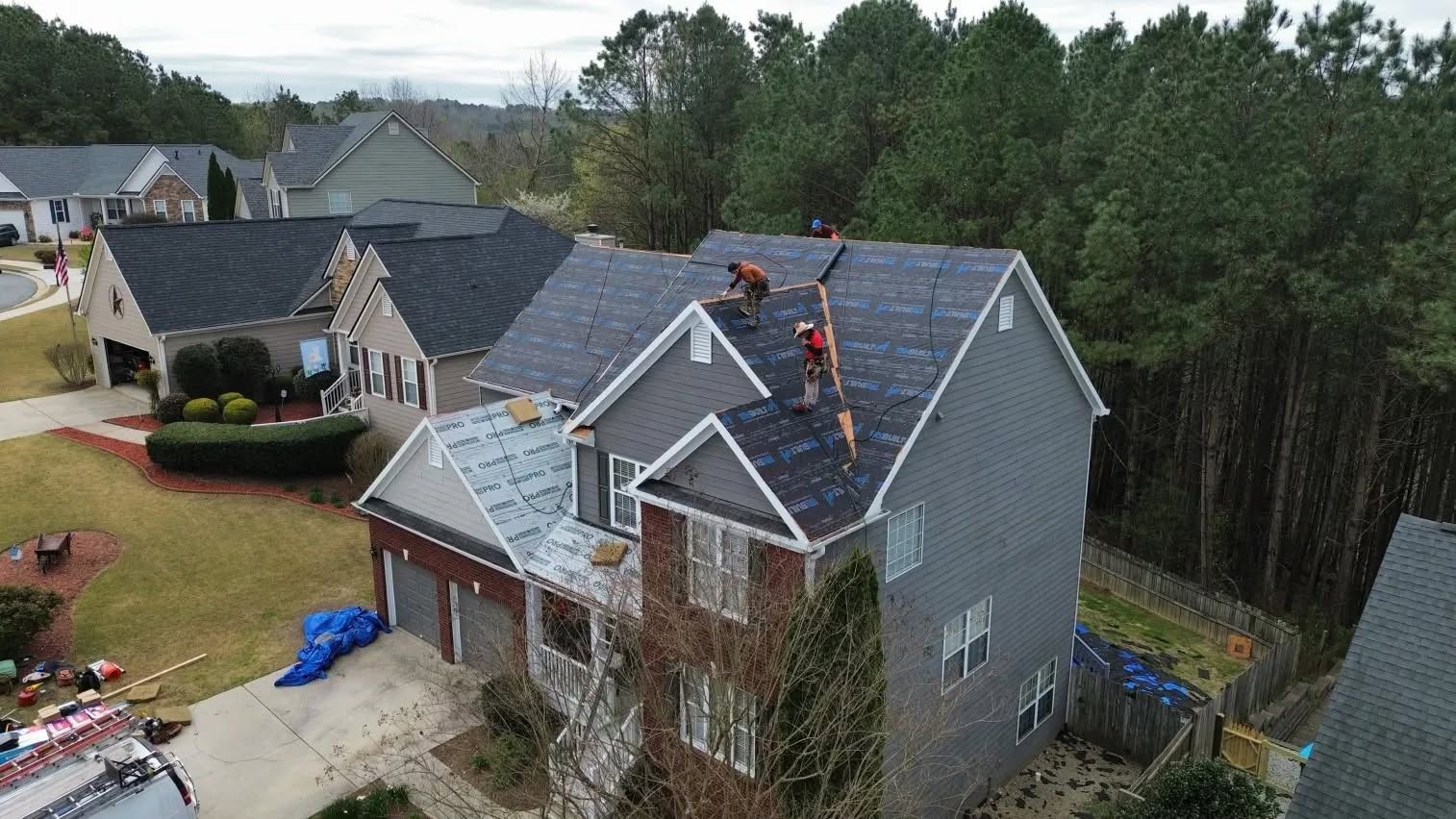 Aerial view of a suburban house with workers on the roof and surrounding trees and neighboring homes