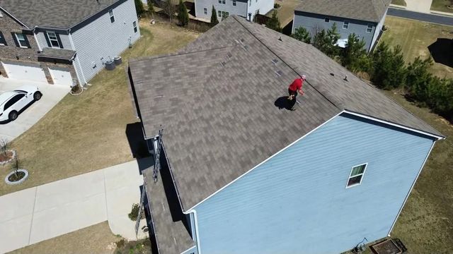 Aerial view of a roof with a person in red working near the ridge on a blue-sided house.