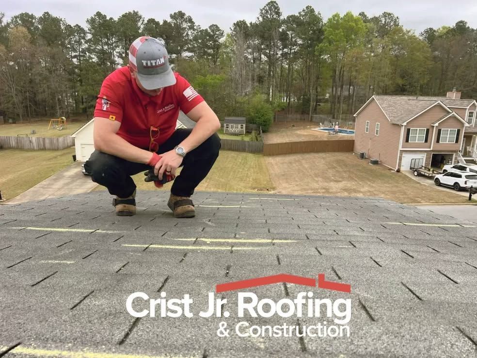 Roofing worker in red shirt crouches on shingled roof, inspecting shingles near suburban homes.