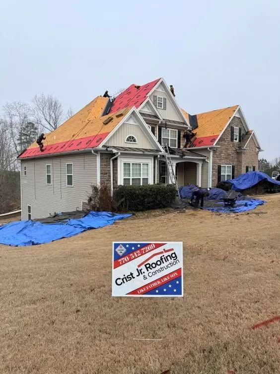 Damaged house with new roof under repair, blue tarps on the lawn, and a C&D Roofing sign in front.