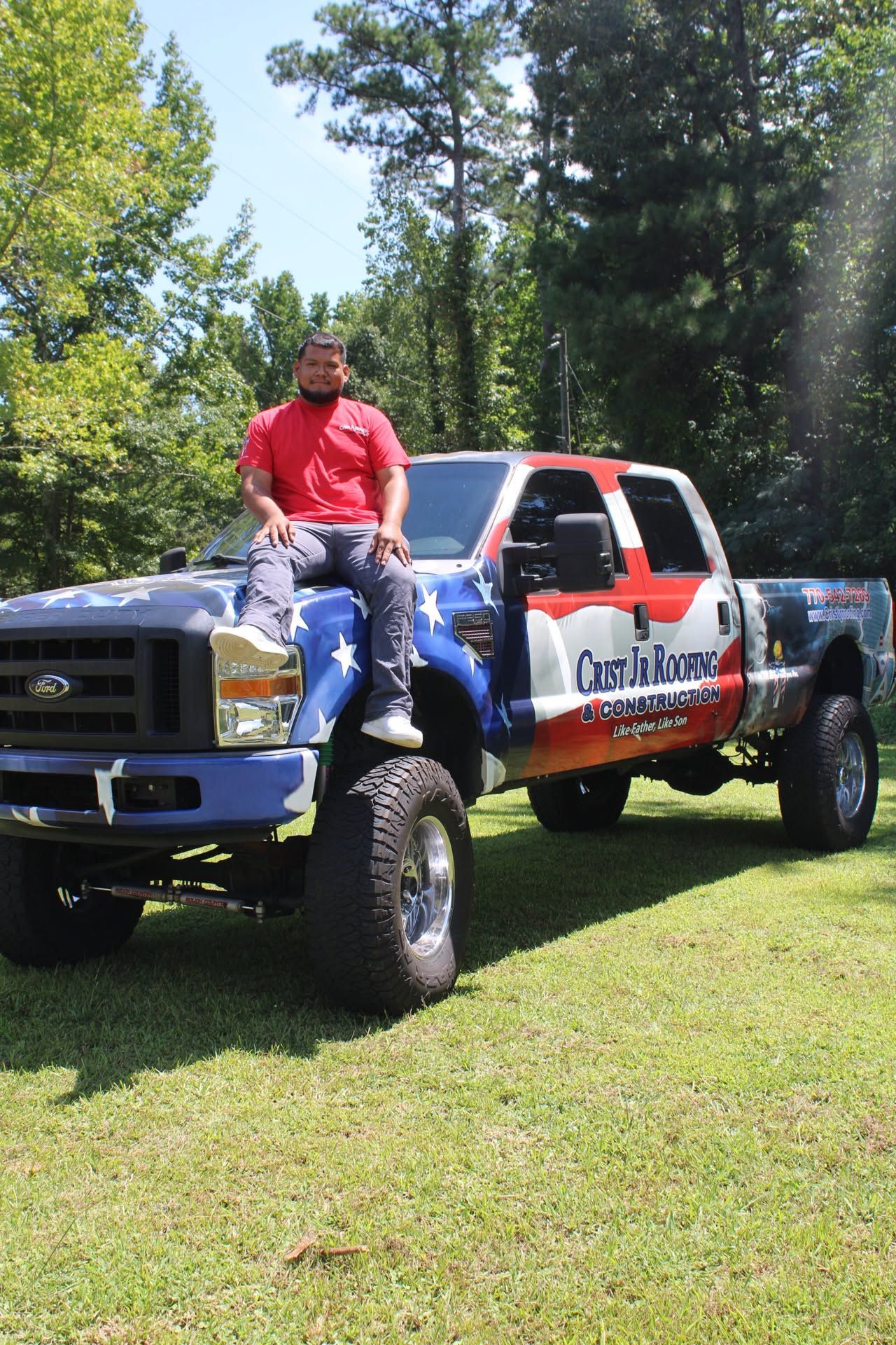 Man sitting on a lifted red-and-white truck in a grassy area with trees in the background