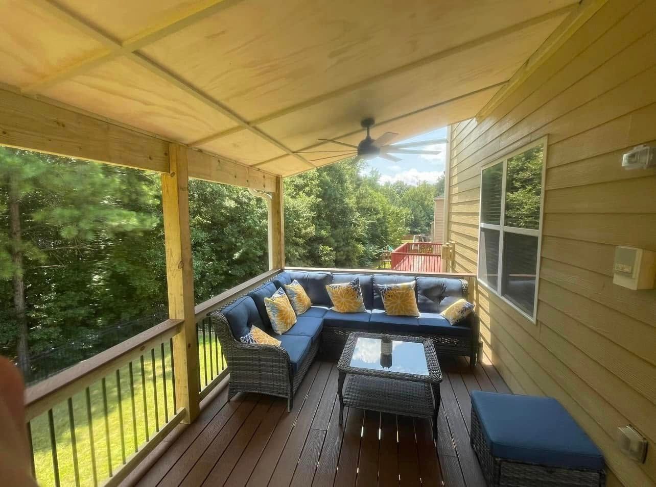 Covered porch with wicker seating, glass coffee table, blue cushions, and wooded view beyond the railing