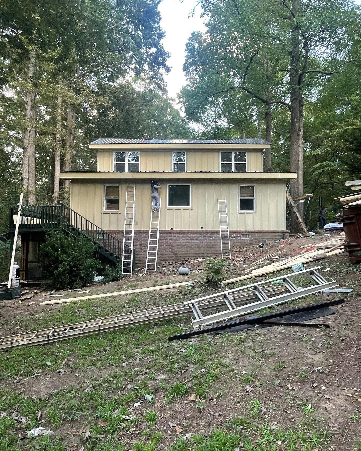 House with ladders and a fallen extension ladder in a wooded yard, suggesting exterior work in progress