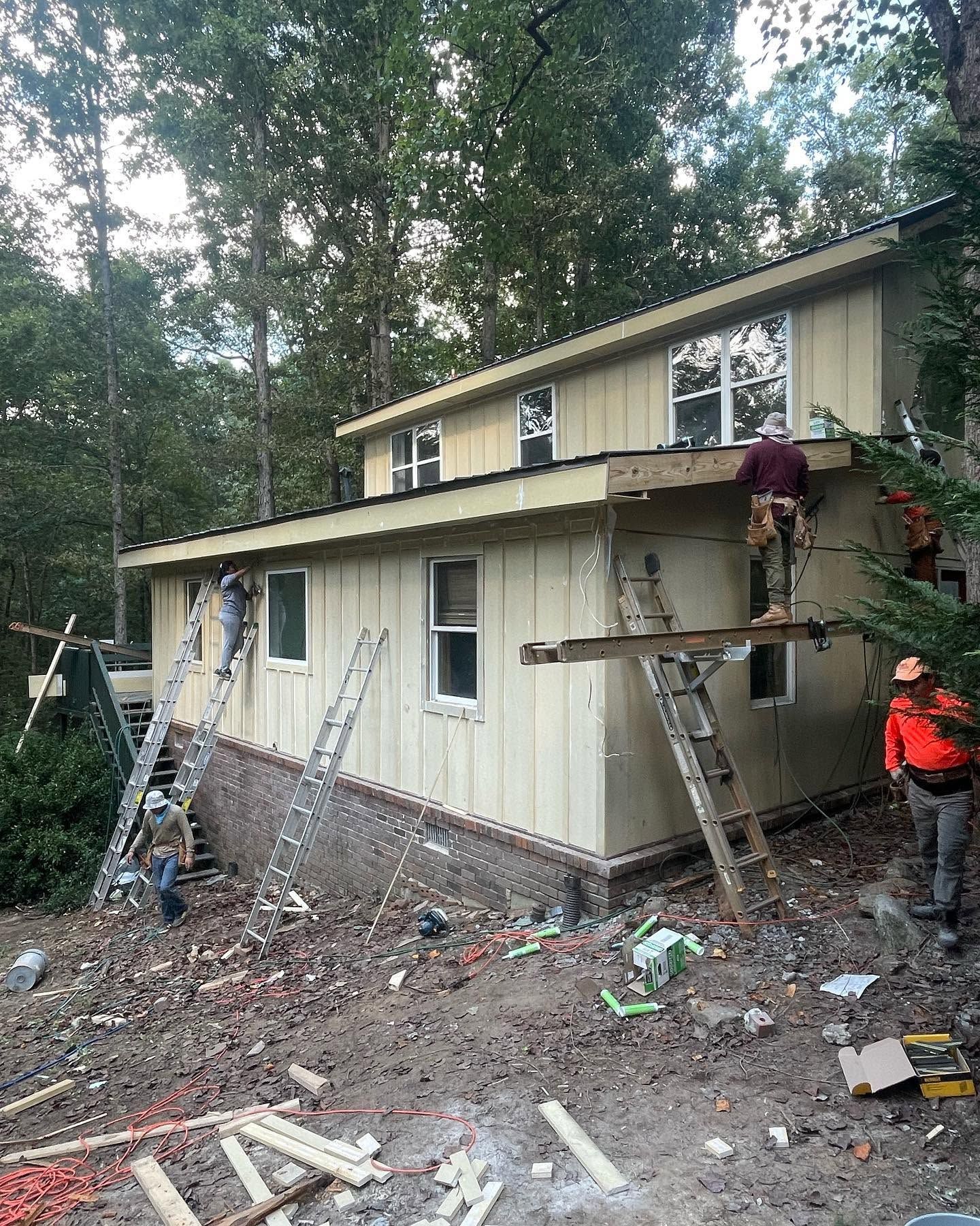 Workers on scaffolding repairing the exterior of a beige two-story house in a wooded area.