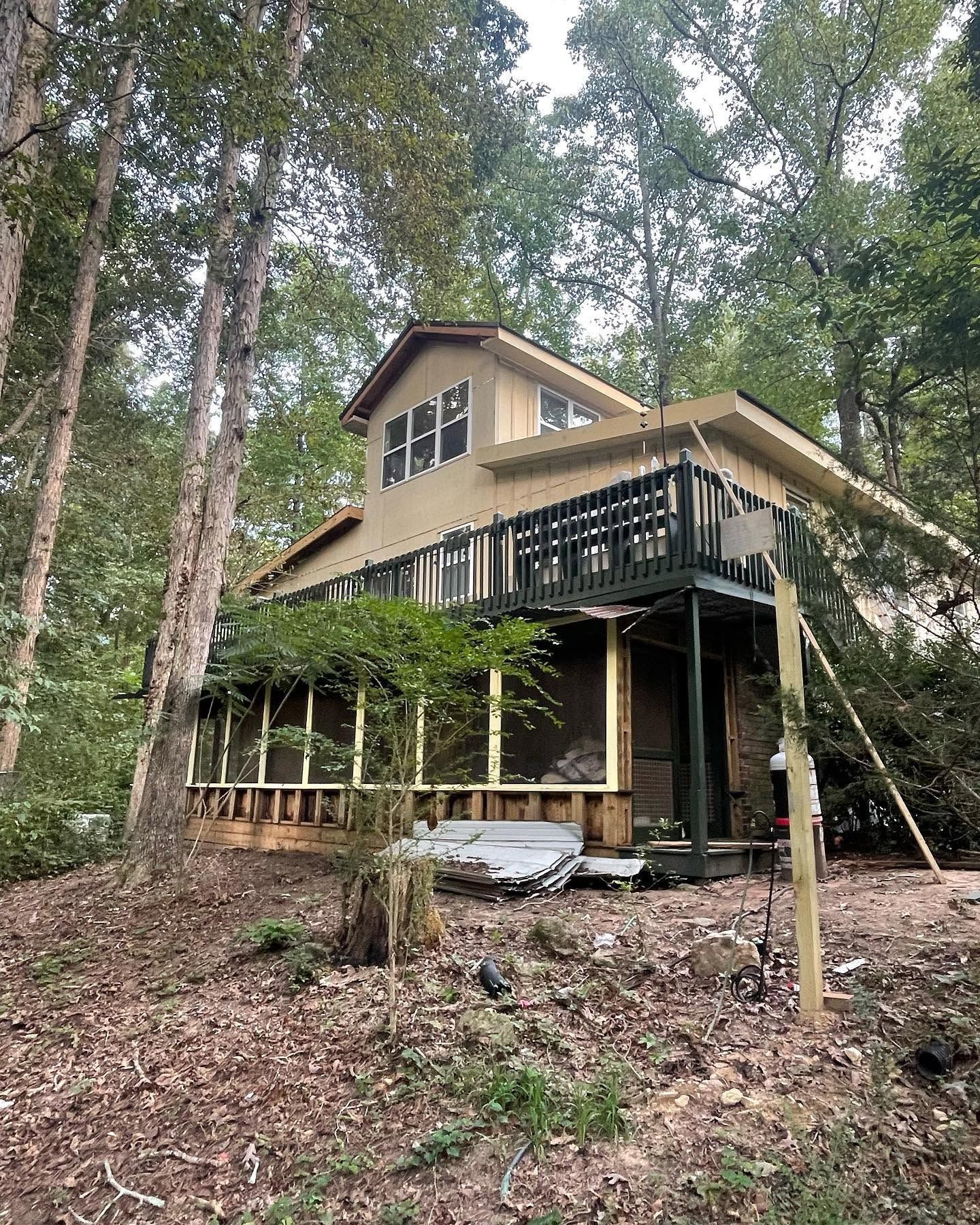 Two-story cabin in the woods with a dark deck and beige siding, surrounded by tall trees and forest floor.