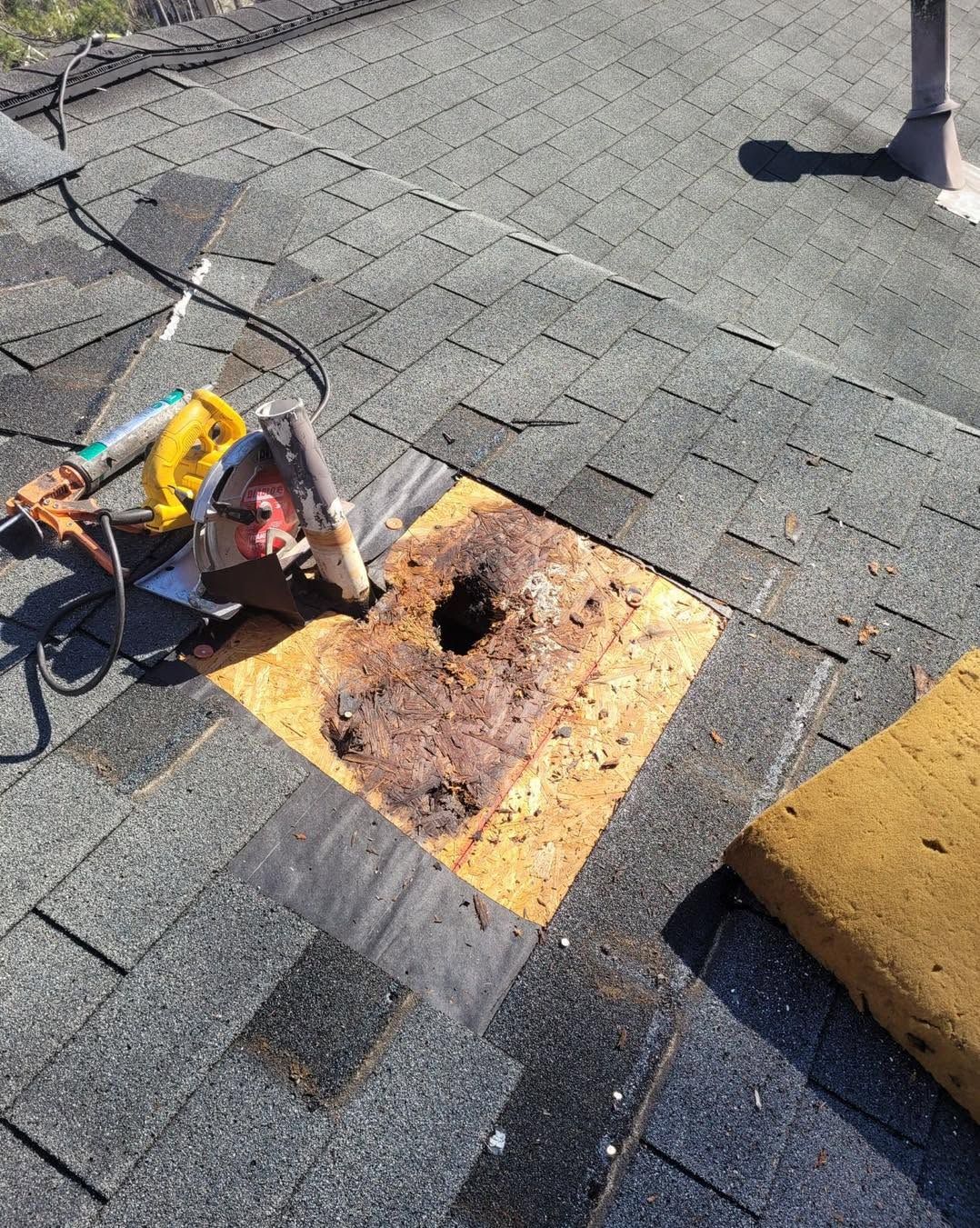 Roof repair tools beside a rusted vent on gray shingles, with a yellow patch and curb block nearby