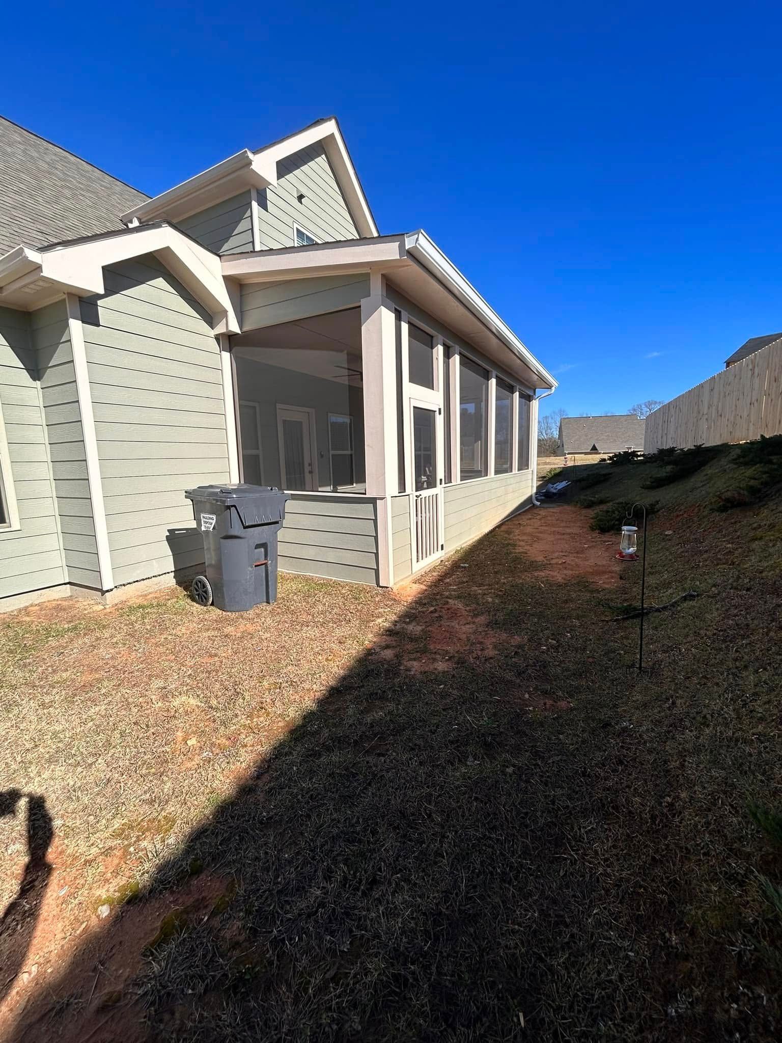 Side view of a light-colored house with a screened porch, trash bins, and a rocky yard under a clear blue sky